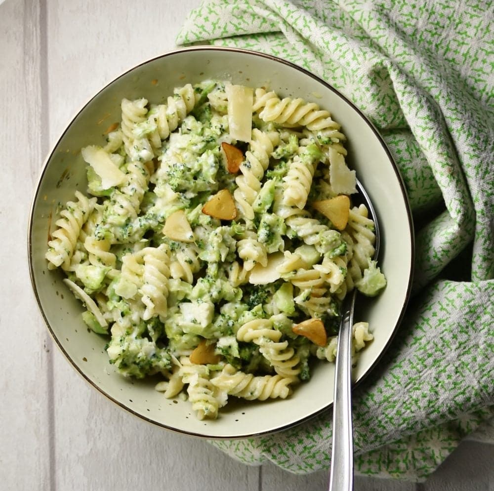 Top down view of pasta with broccoli and spoon in light green coloured bowl wrapped in green cloth.