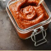 Top down view of barbecue sauce in square open jar with spoon and green cloth in background.