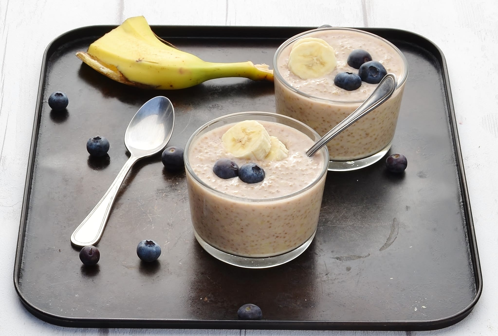 Overnight quinoa with banana slices and blueberries in 2 cups with spoon, another spoon, blueberries and banana peel on dark table.