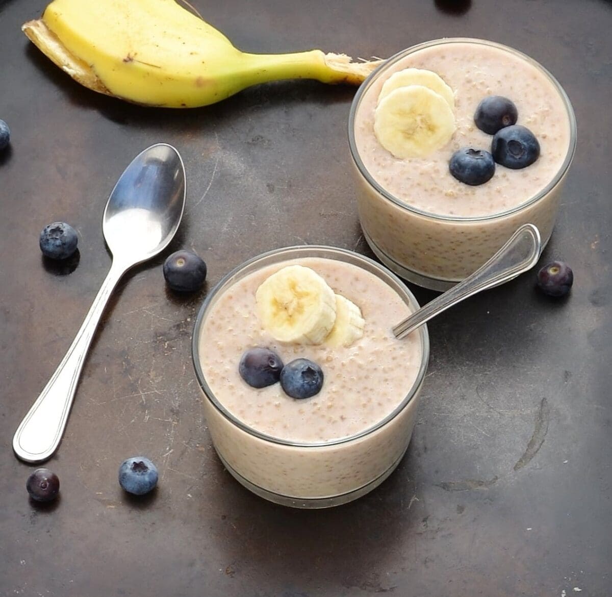 Overnight quinoa with banana slices and blueberries in 2 cups with spoon, another spoon, blueberries and banana peel on dark table.