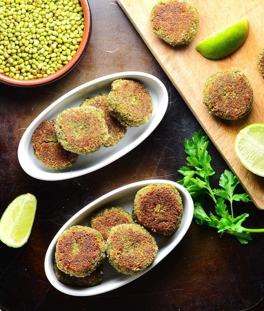 Top down view of bean and mushroom patties with lime, beans in brown dish and cutting board on oven tray.