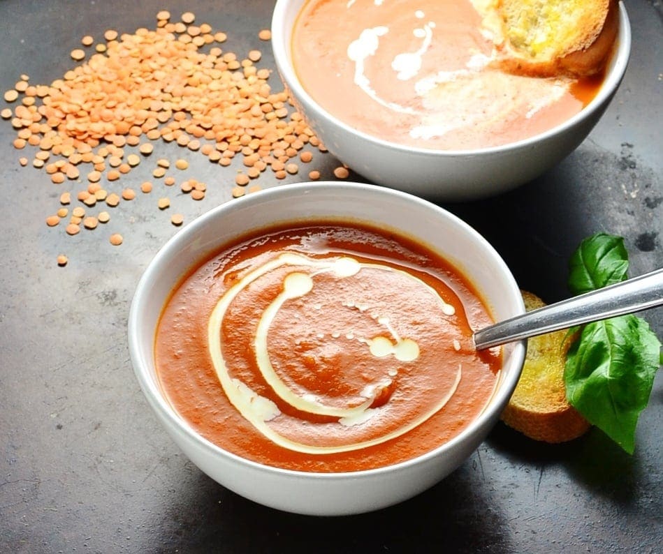 Side view of tomato and lentil soup in white bowls with spoon, red lentils, basil and toast on black background.