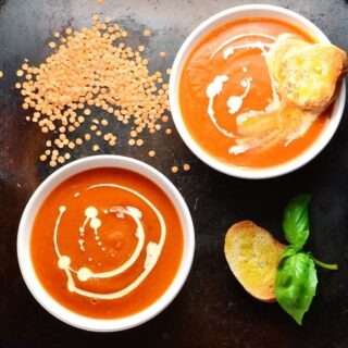 Top down view of tomato and lentil soup in 2 white bowls with toasted bread, lentils and basil leaves on oven tray.