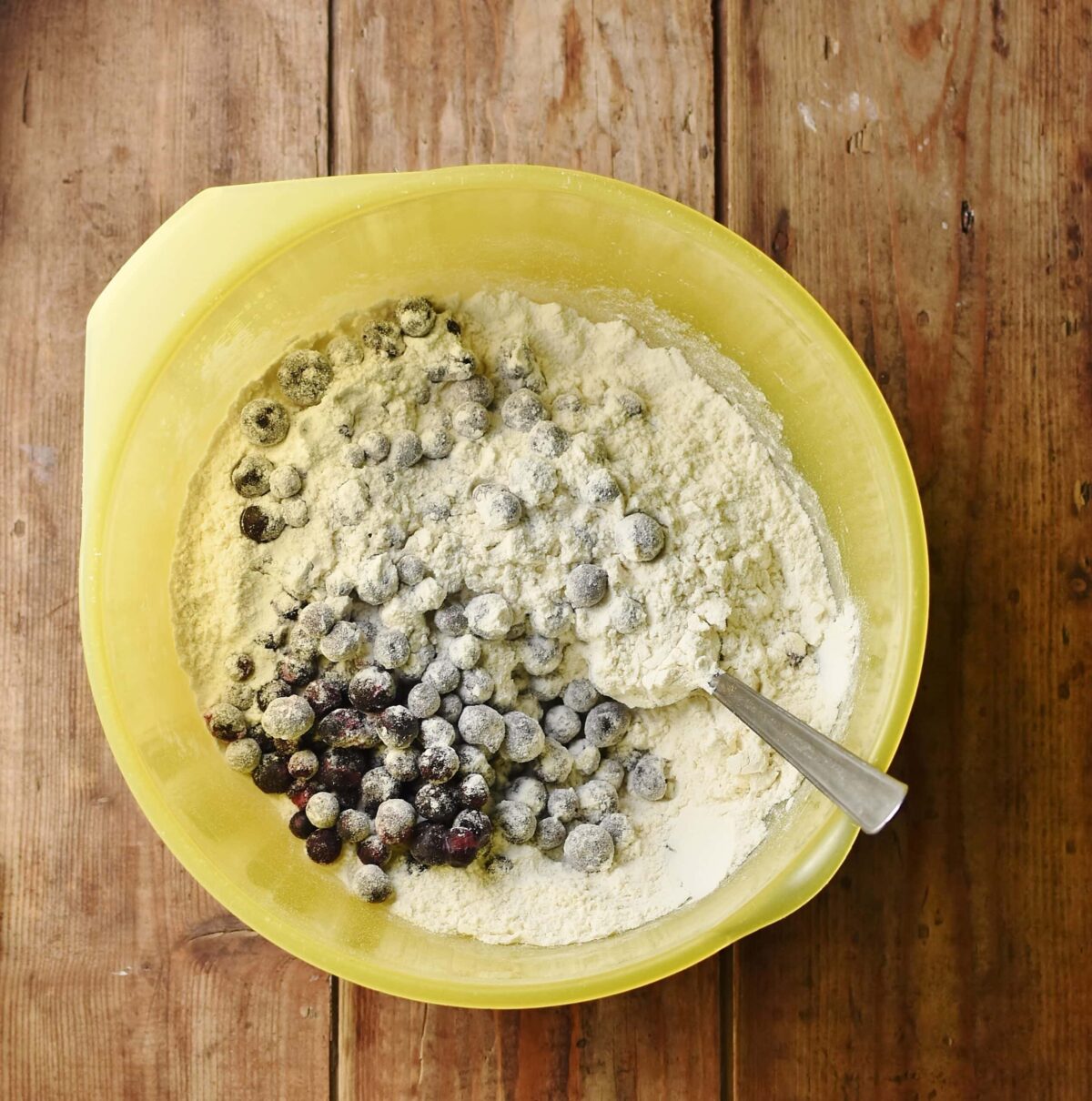 Blueberries and flour mixture in large yellow bowl with spoon.