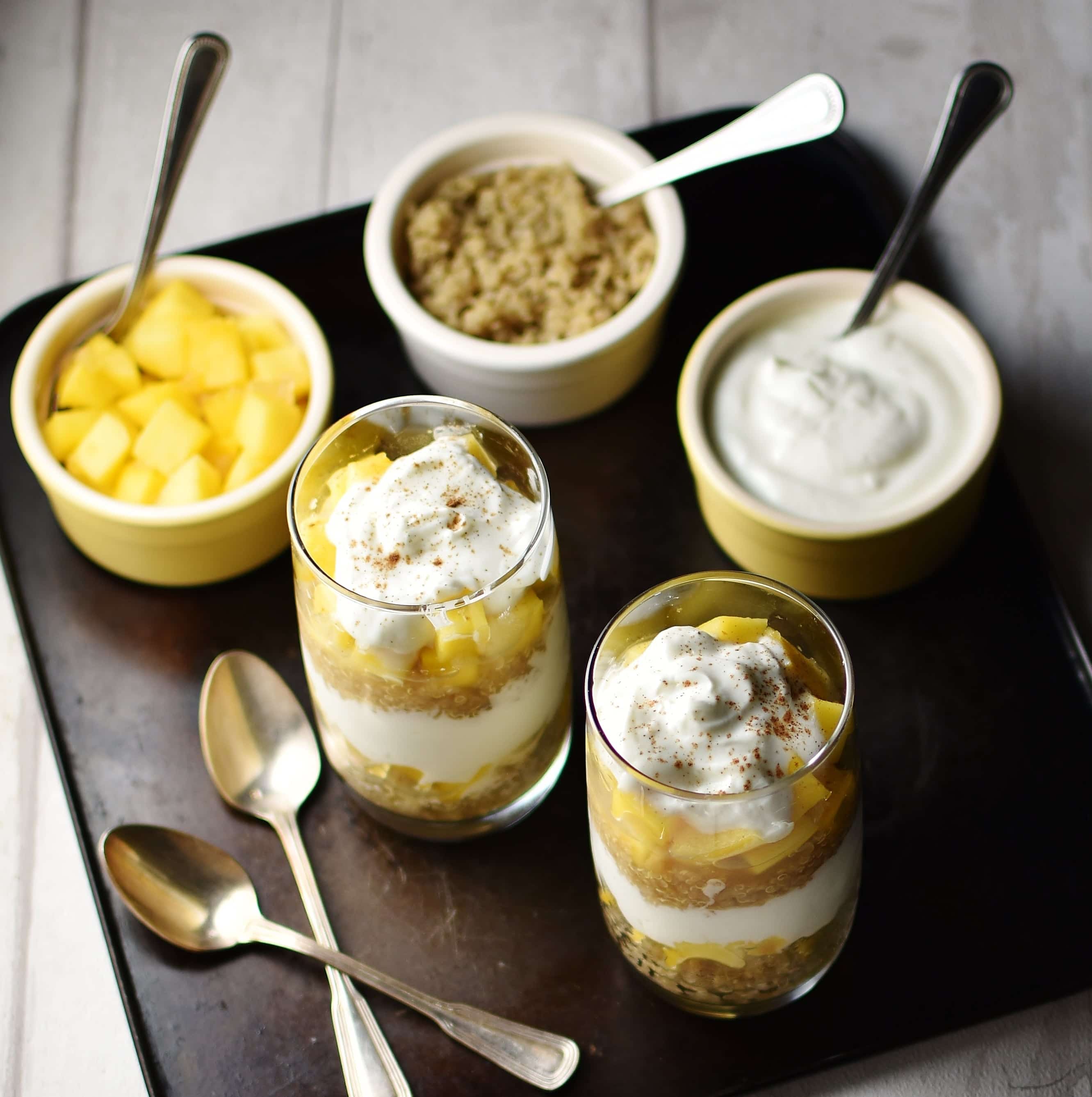 Yogurt, mango, quinoa parfait in 2 tall glasses, 2 spoons and 3 dishes with mango cubes, quinoa and yogurt, with spoons, in background on top of dark tray.