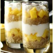 Side view of yogurt, mango, quinoa parfait in 2 tall glasses, with spoons on top of dark tray.