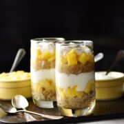 Side view of yogurt, mango, quinoa parfait in 2 tall glasses, 2 spoons and 2 yellow dishes with spoons in background on top of dark tray.