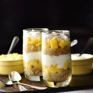 Side view of yogurt, mango, quinoa parfait in 2 tall glasses, 2 spoons and 2 yellow dishes with spoons in background on top of dark tray.