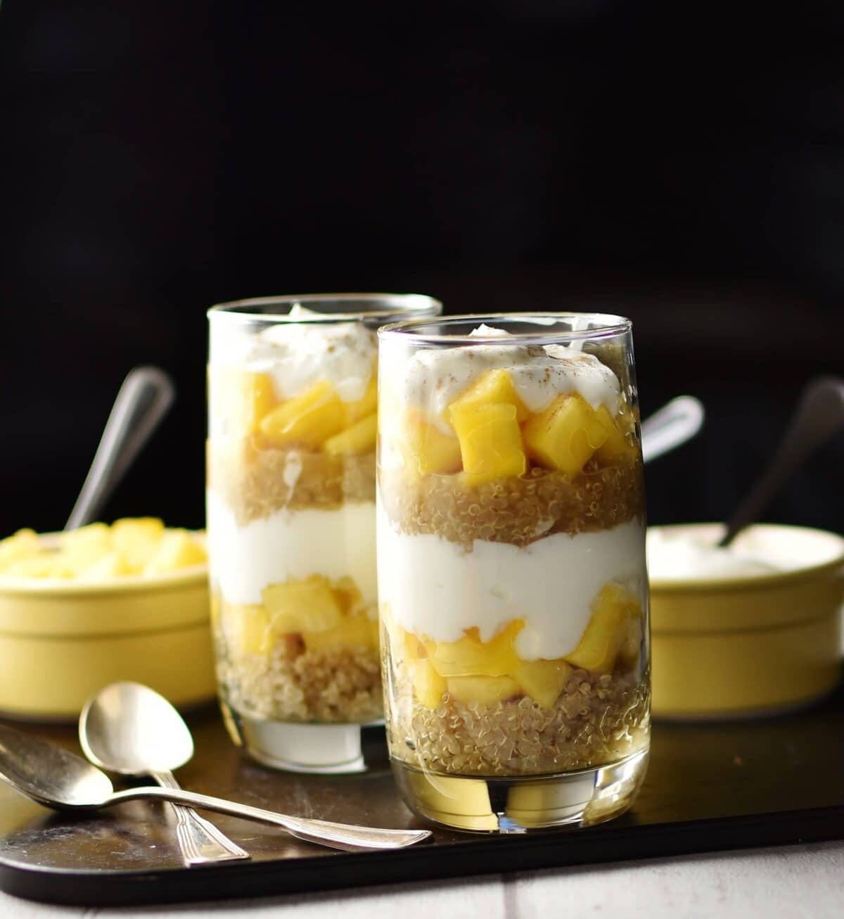 Side view of yogurt, mango, quinoa parfait in 2 tall glasses, 2 spoons and 2 yellow dishes with spoons in background on top of dark tray.