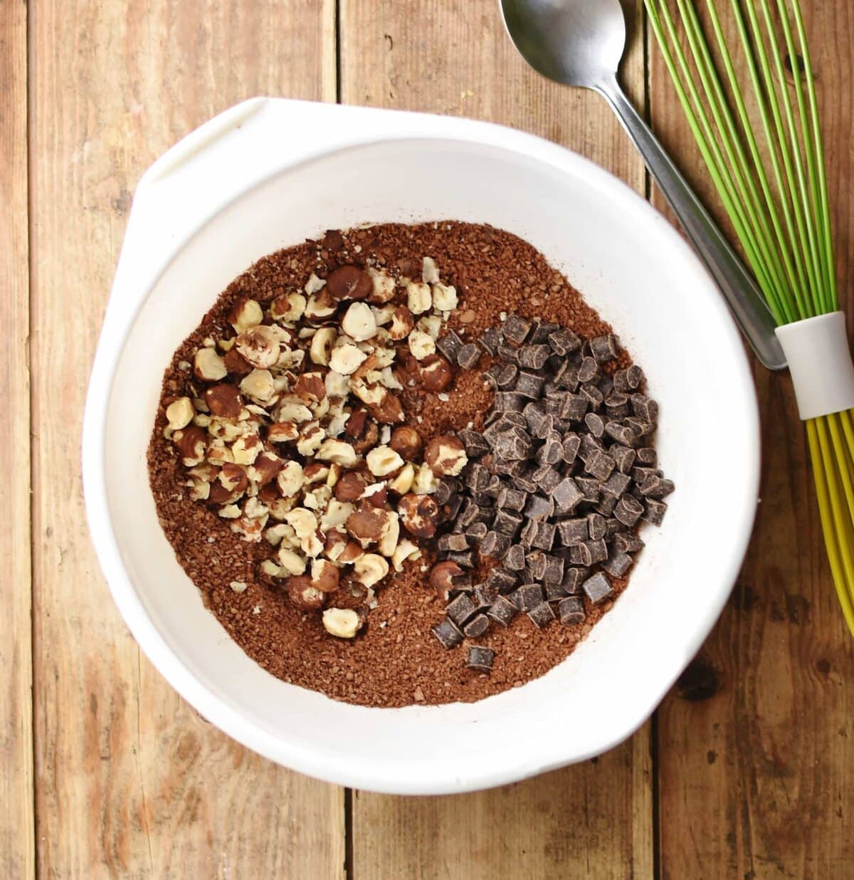Cocoa mixture with crushed nuts and chocolate chunks inside large white bowl with spoon and green whisk in top right.