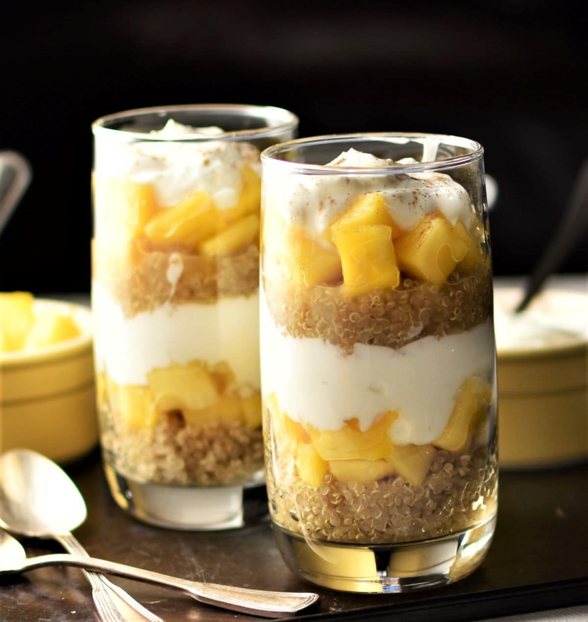 Close-up side view of quinoa yogurt breakfast parfait in 2 tall glasses with spoons in background.