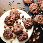 Top down view of almond flour chocolate cookies on white plate with hazelnuts and chocolate chunks on oven tray.