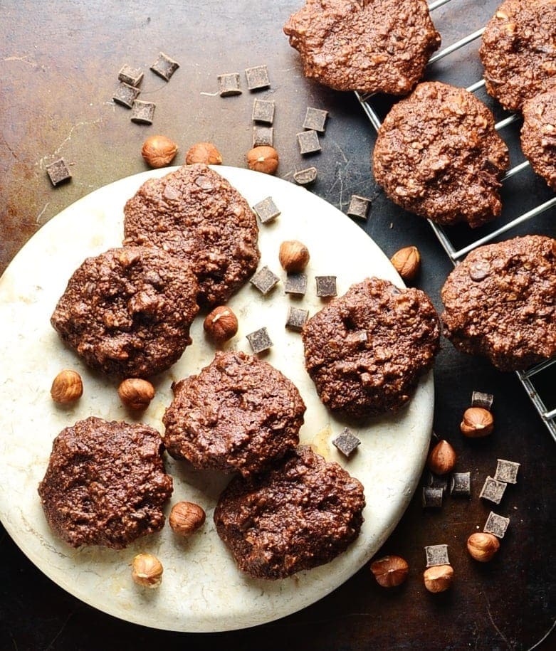 Top down view of chocolate cookies on white plate with hazelnuts and chocolate chunks on oven tray.