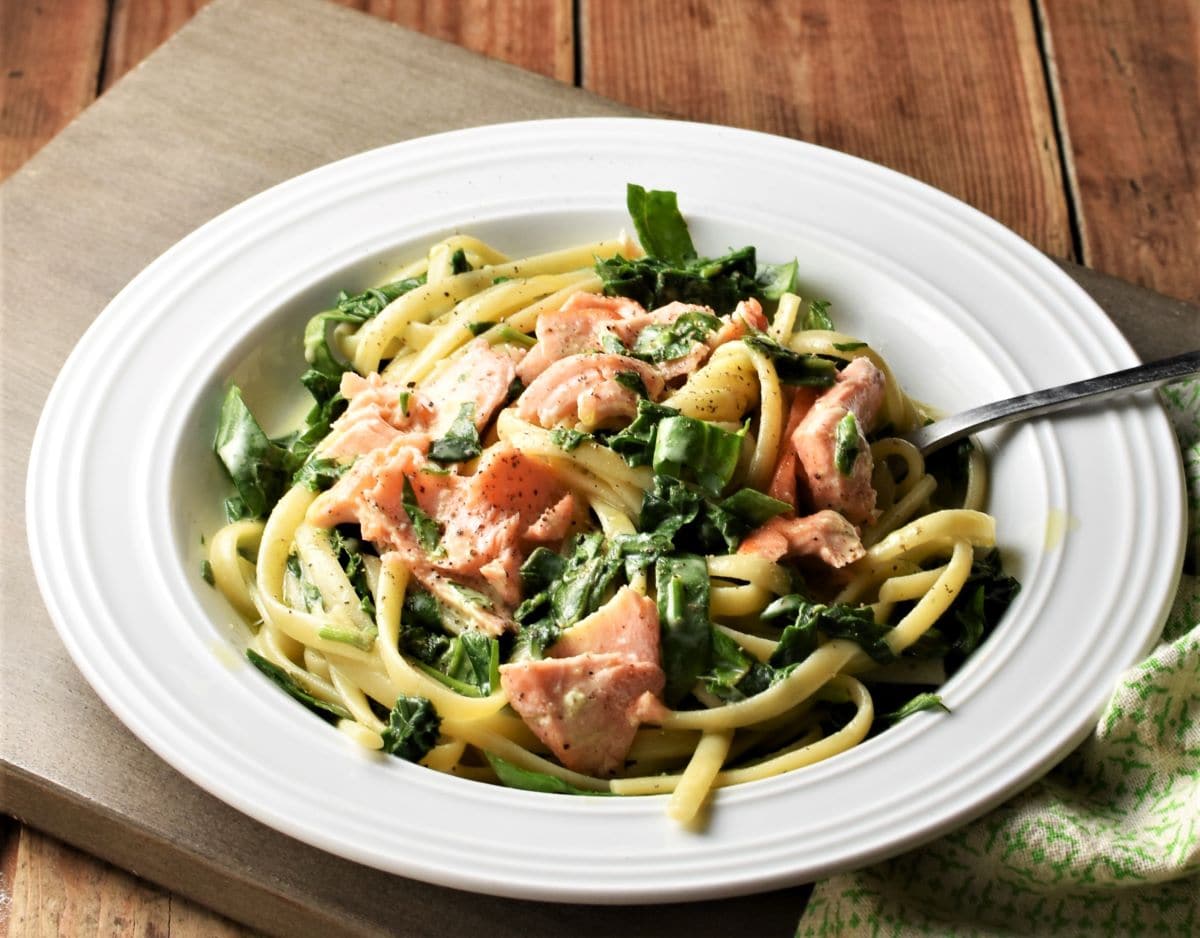 Side view of pasta with spinach and salmon in white bowl with fork.
