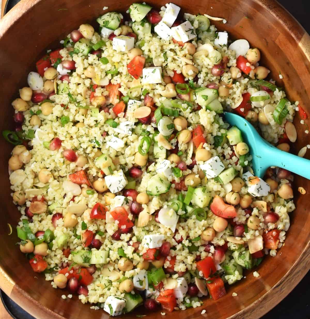 Bulgur wheat salad with chickpeas, vegetables, pomegranate, feta and herbs in large wooden bowl with blue spoon.