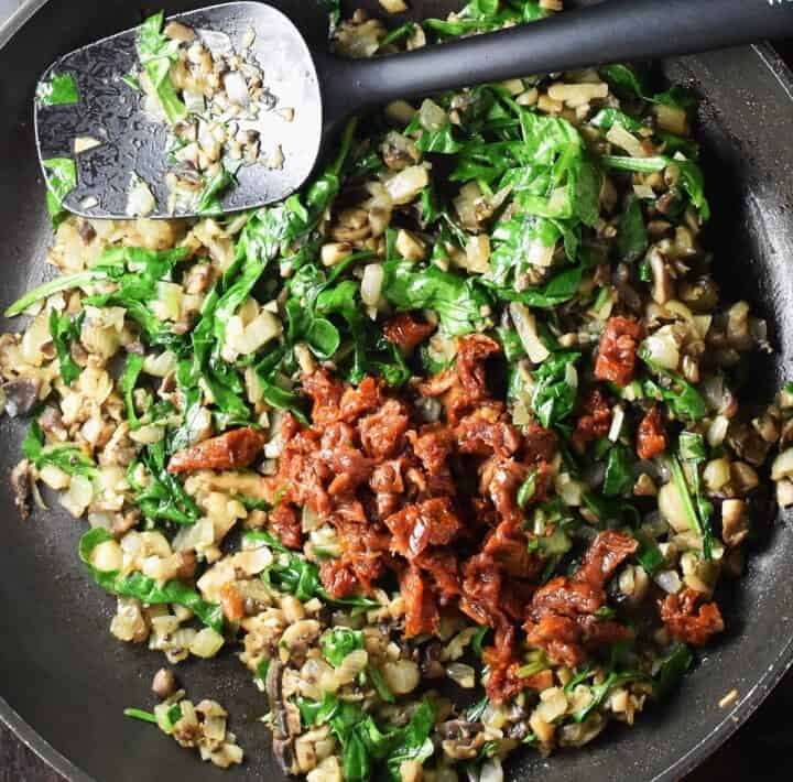 Mushrooms with spinach and tomato in large pan with black spoon.