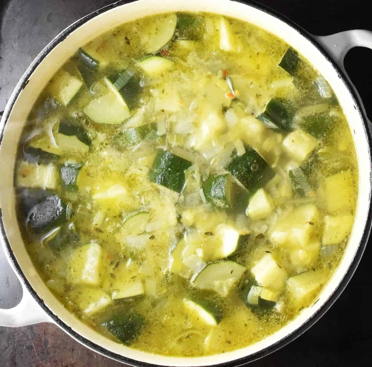 Zucchini and potatoes cooking in stock for soup in white pot.