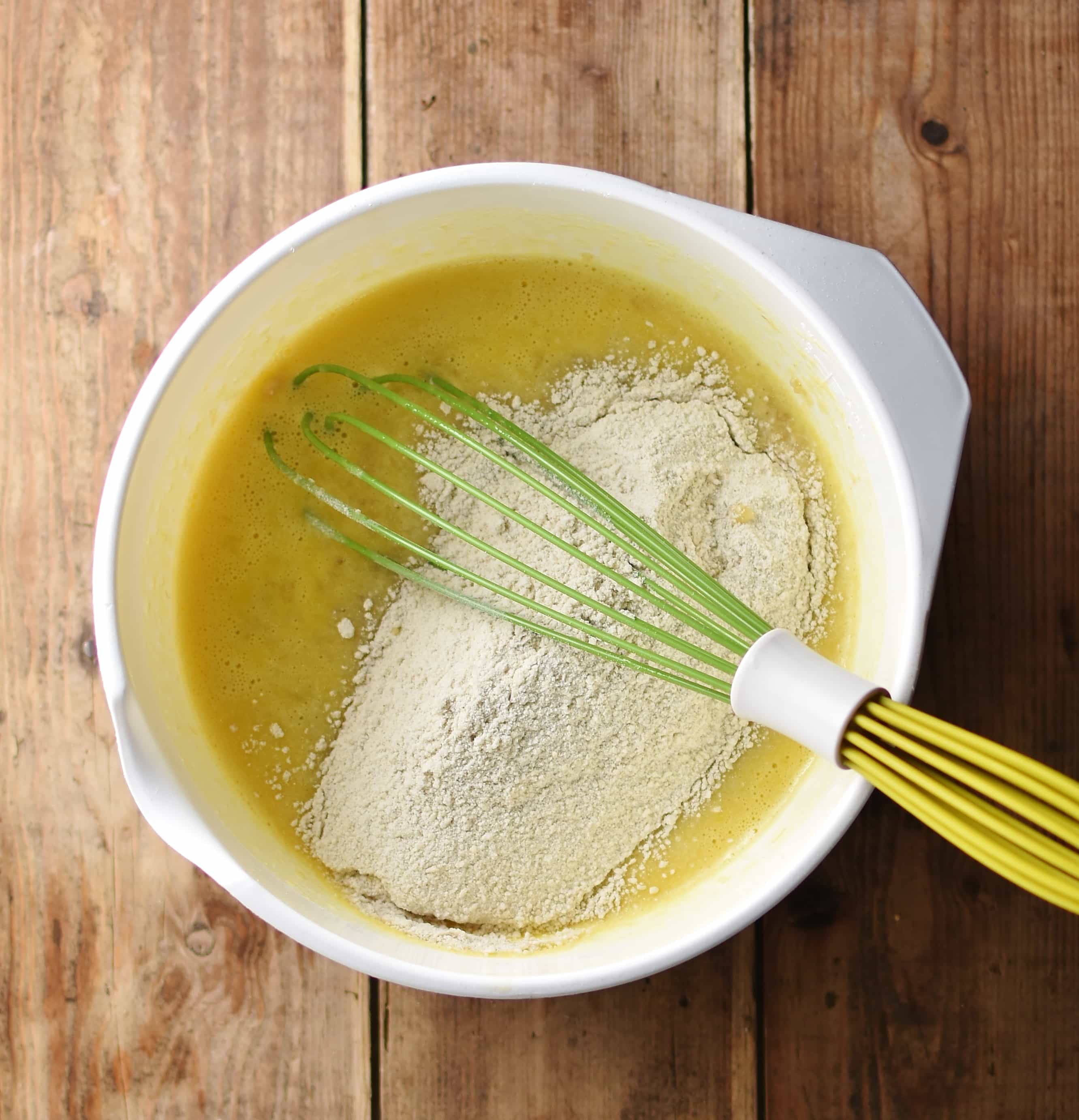 Wet and dry banana bread ingredients in large white bowl with green whisk.