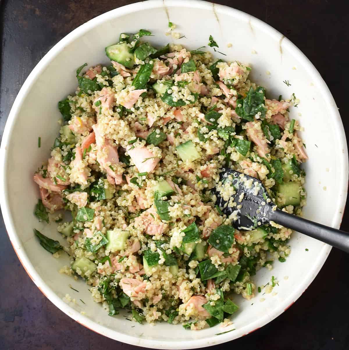 Top down view of assembled quinoa and salmon salad with cucumber and spinach in white bowl with spoon.