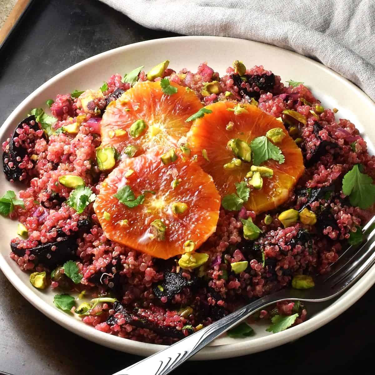 Side view of salad with quinoa, roasted beets and orange slices on top on pink plate with fork.