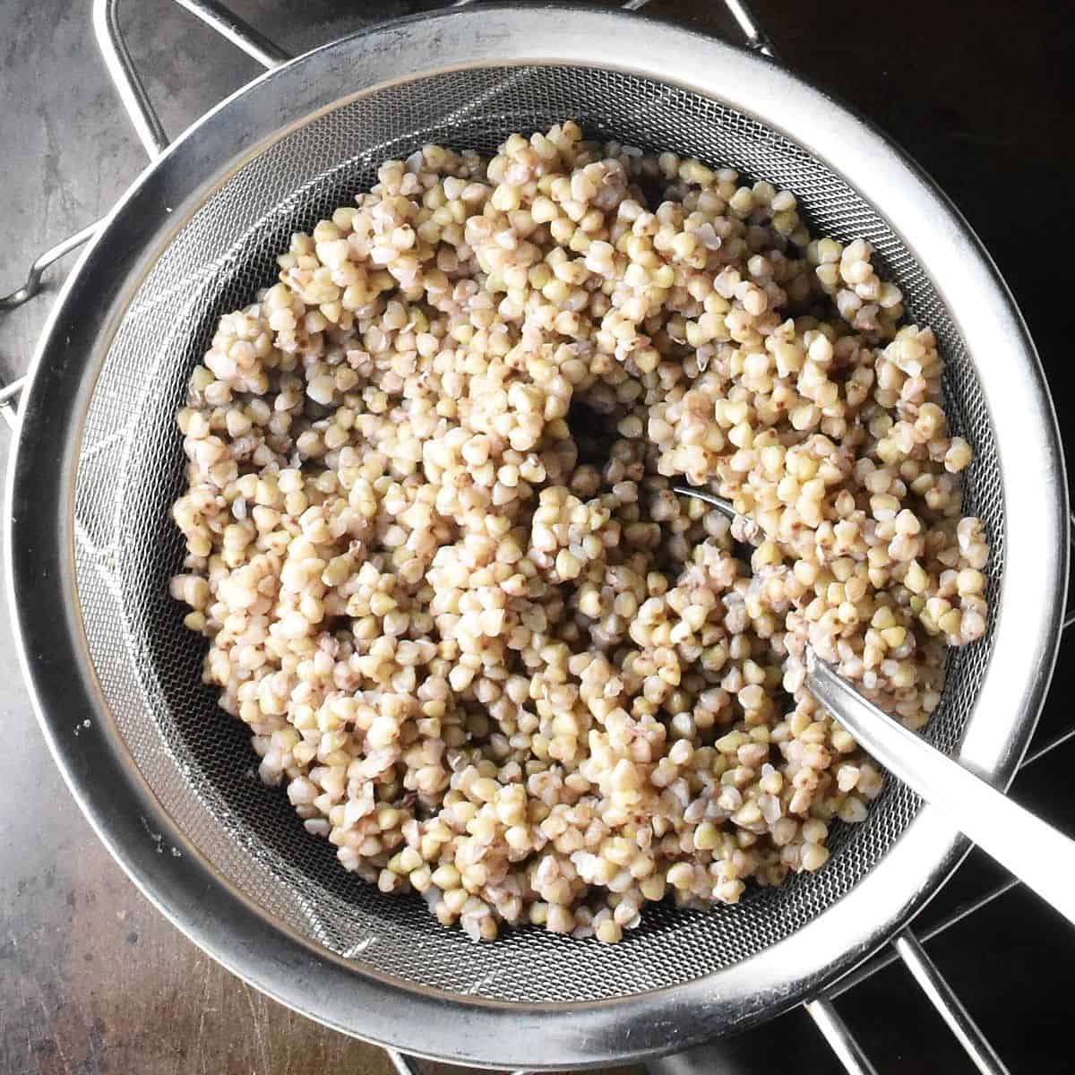 Top down view of cooked buckwheat groats in metal strainer with spoon.