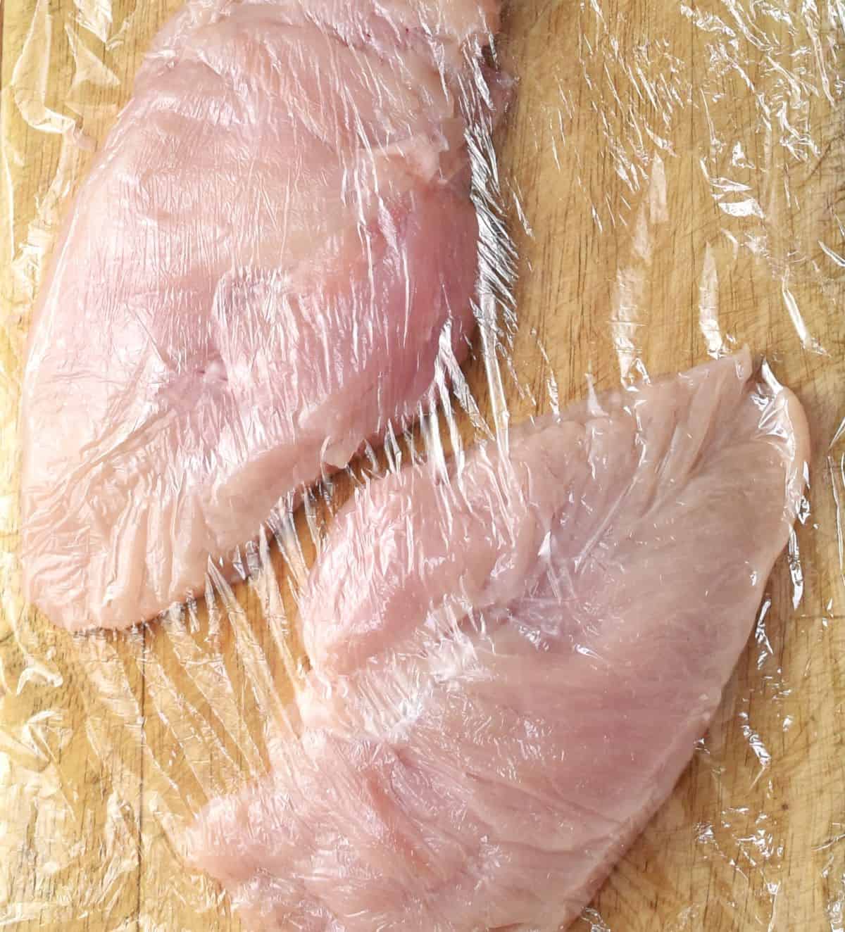2 pieces of chicken covered with cling wrap on cutting board.