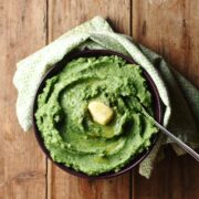 Top down view of mashed potatoes with spinach and knob of butter in bowl with spoon wrapped in green cloth.