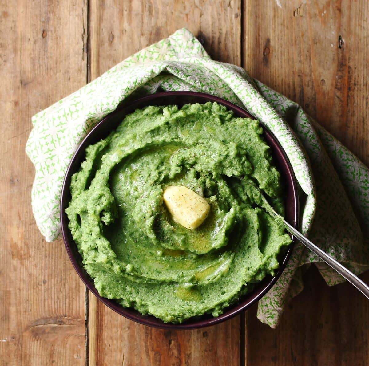 Top down view of mashed potatoes with spinach and knob of butter in bowl with spoon wrapped in green cloth.