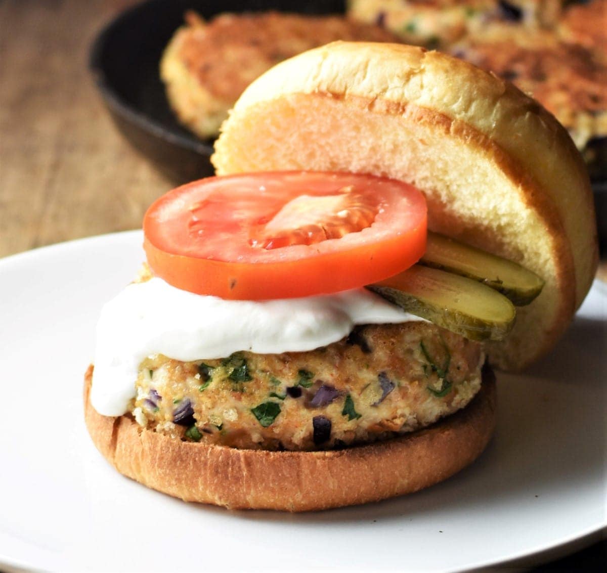 Chicken burger in open bun with sauce, slice of tomato and pickles.