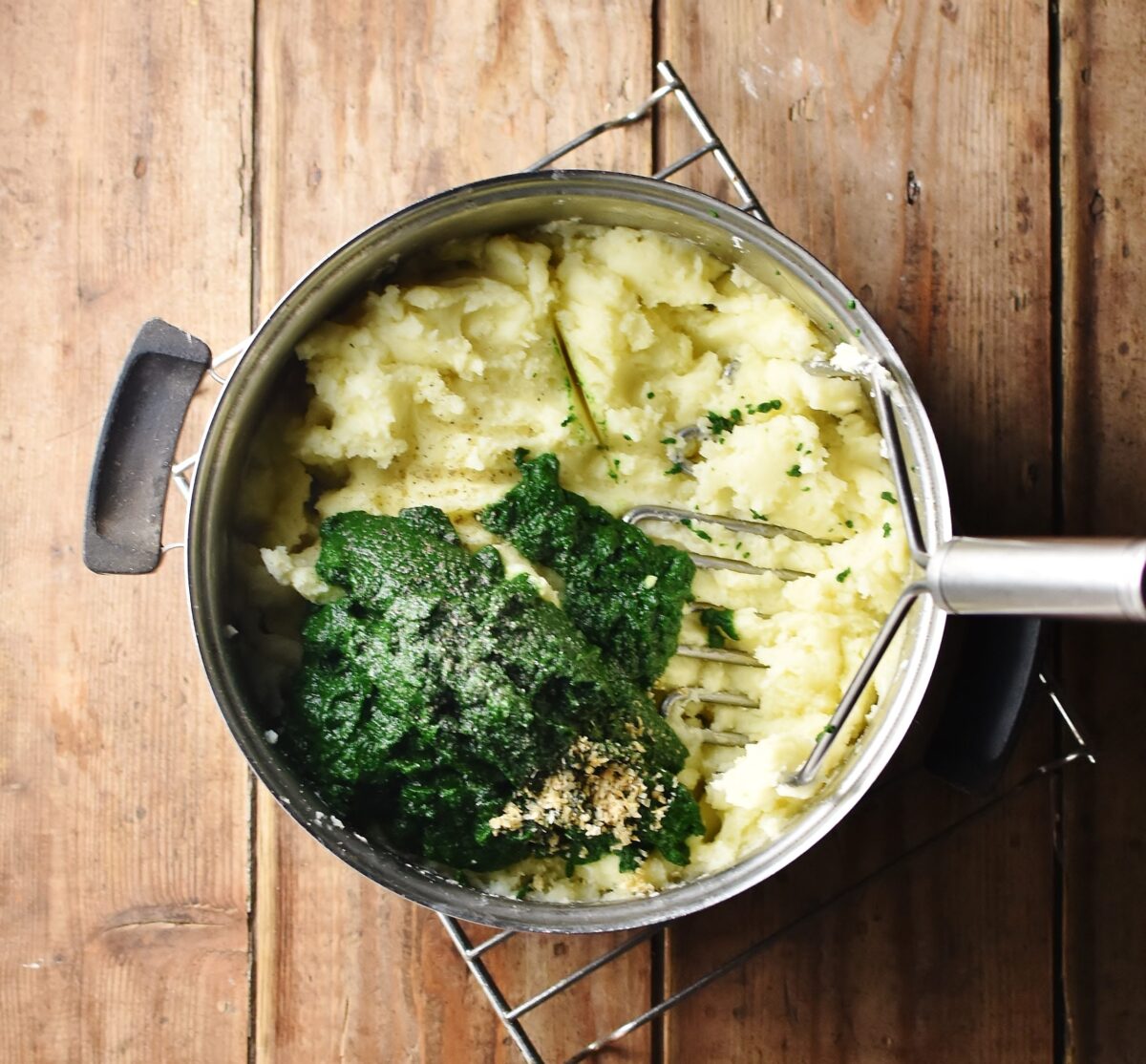 Mashed potatoes with pureed spinach and potato masher in pot.
