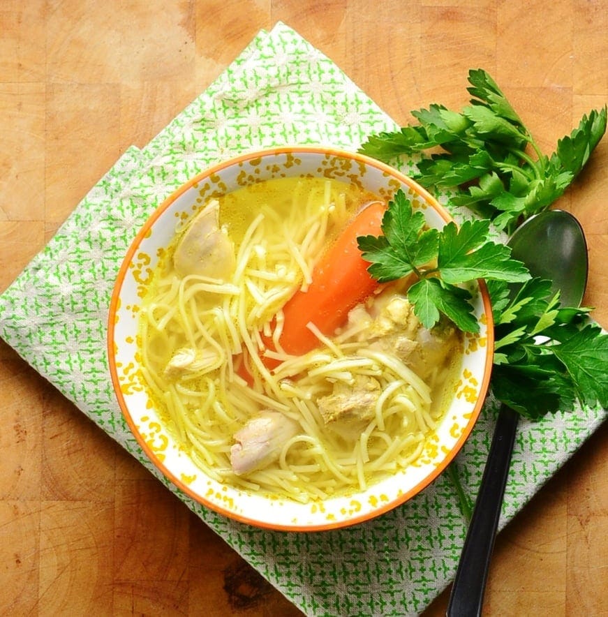 Chicken noodle rosol soup in orange bowl with spoon and parsley on green cloth.
