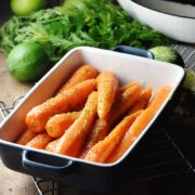 Side view of braised carrots in rectangular blue dish, with limes, carrot tops and white pan in background.