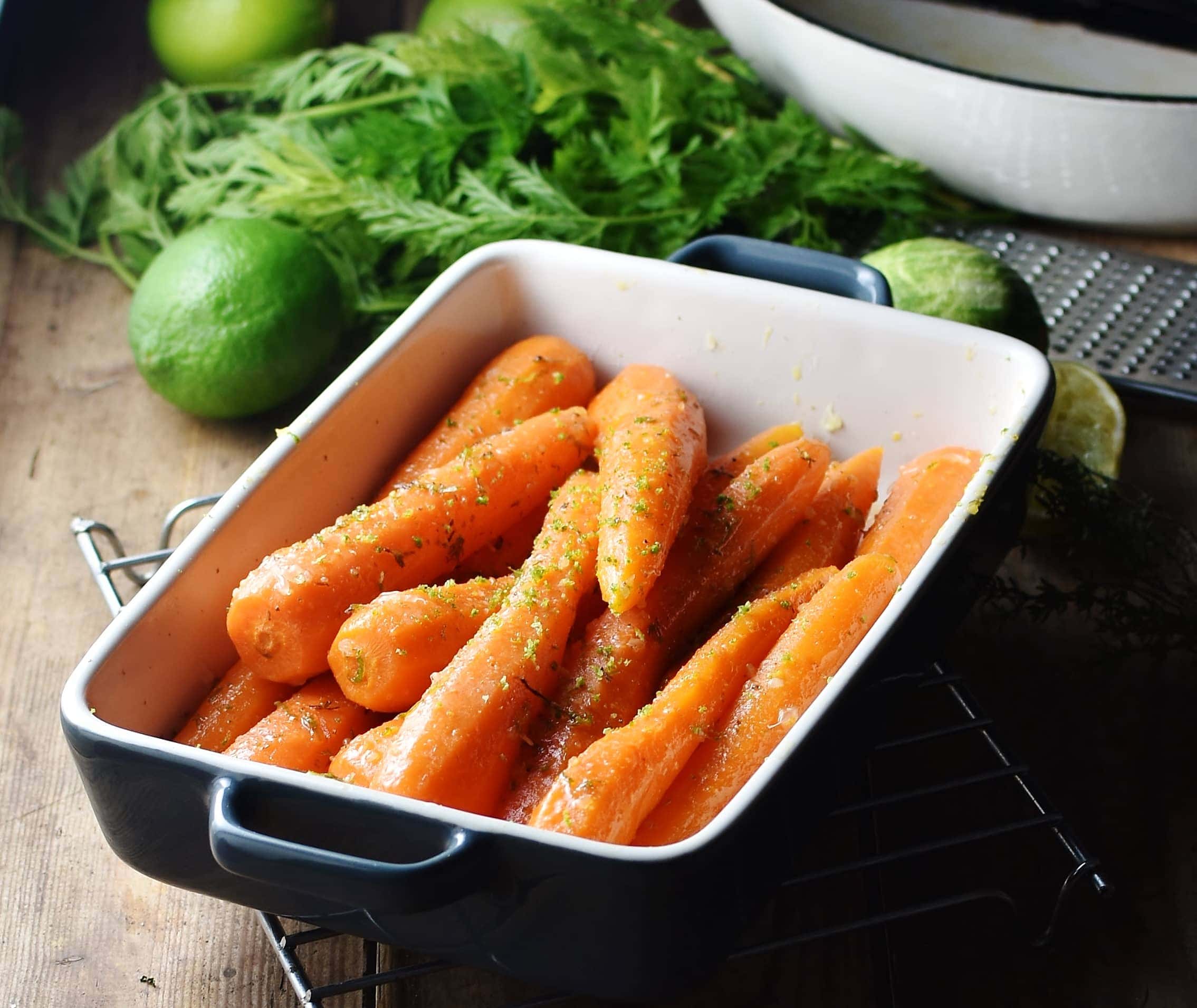 Side view of braised carrots in rectangular blue dish, with limes, carrot tops and white pan in background.