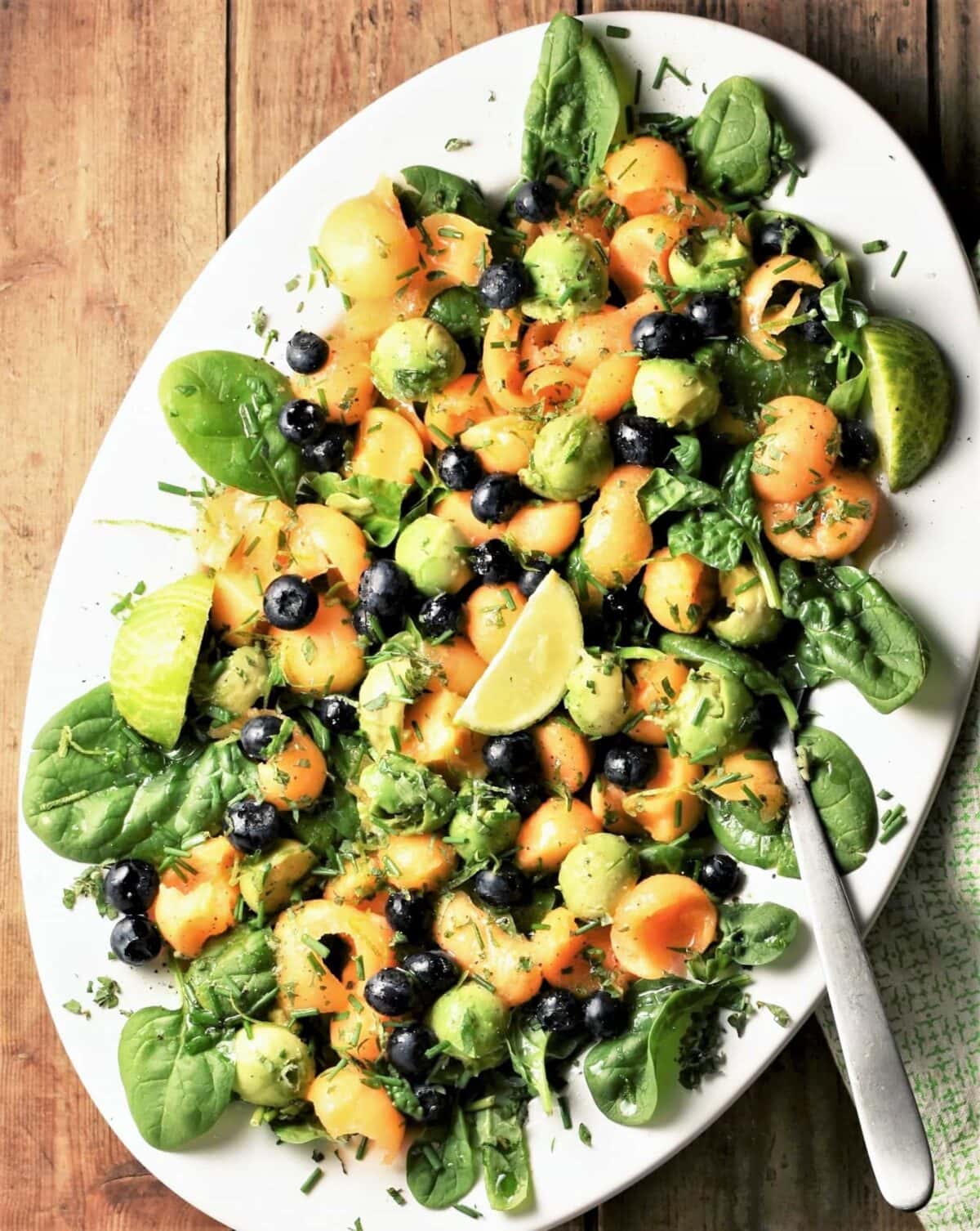Top down view of cantaloupe salad with spinach and avocado on white oval plate with spoon.