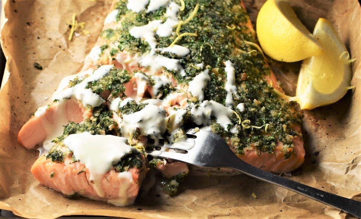 Close-up partial view of herb crusted salmon with creamy sauce, fork and lemon wedges.