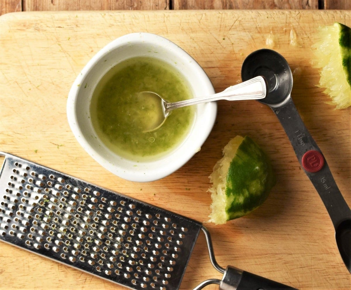 Lime dressing in white bowl with spoon, lime and zester in background.