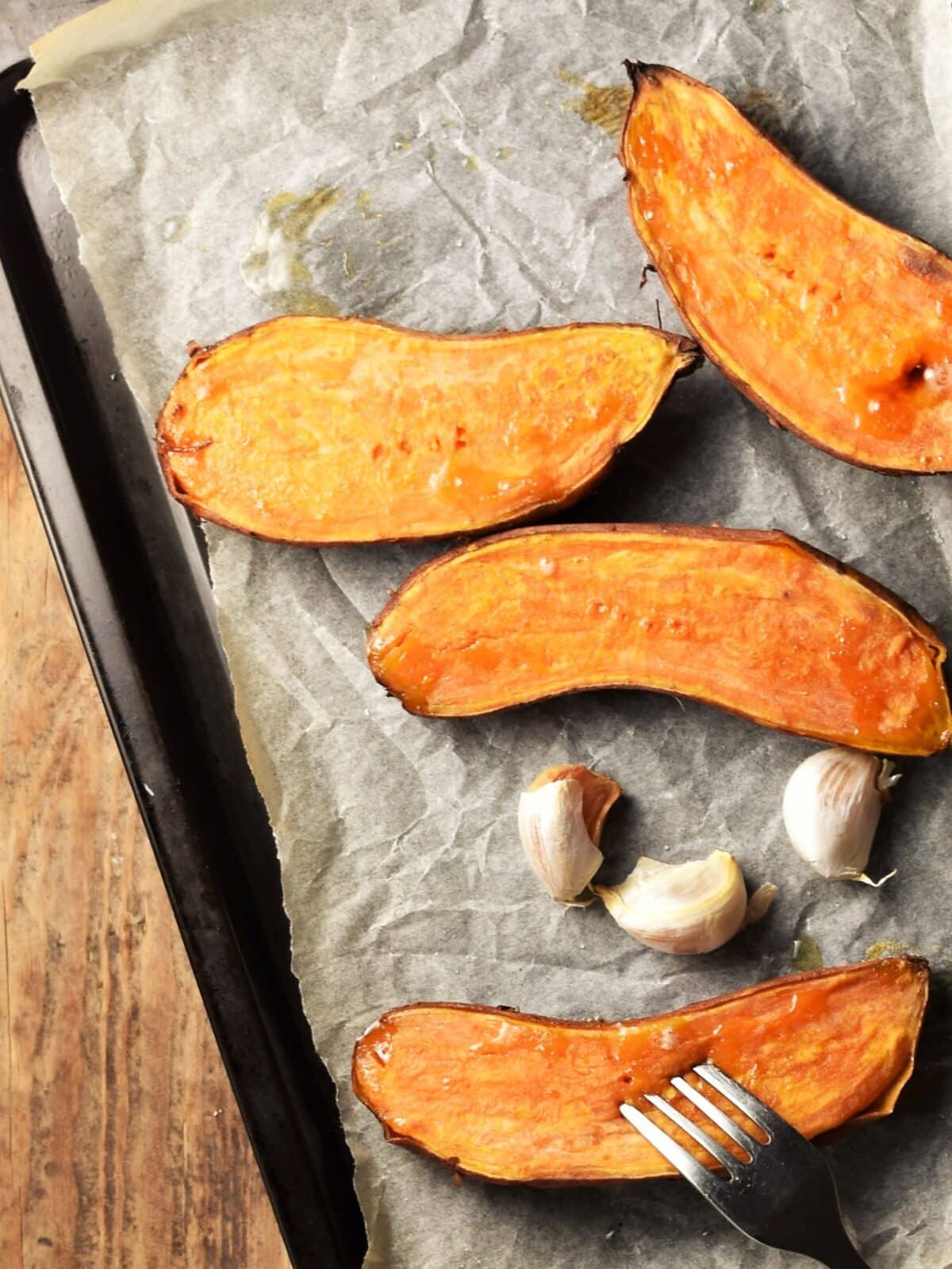 Top down view of halved roasted sweet potatoes and garlic cloves on tray lined with parchment paper.