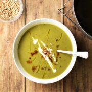 Green sweet potato soup with seeds, drizzle of yogurt and balsamic vinegar in white bowl with spoon, and sunflower seeds in small dish in top left corner.