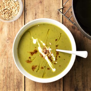 Green sweet potato soup with seeds, drizzle of yogurt and balsamic vinegar in white bowl with spoon, and sunflower seeds in small dish in top left corner.