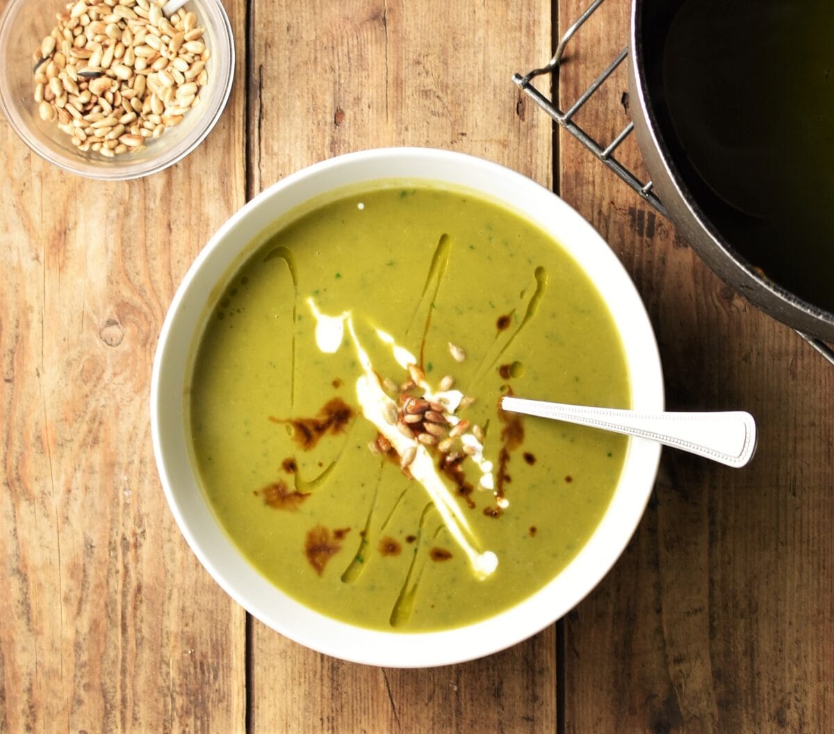 Green sweet potato soup with seeds, drizzle of yogurt and balsamic vinegar in white bowl with spoon, and sunflower seeds in small dish in top left corner.