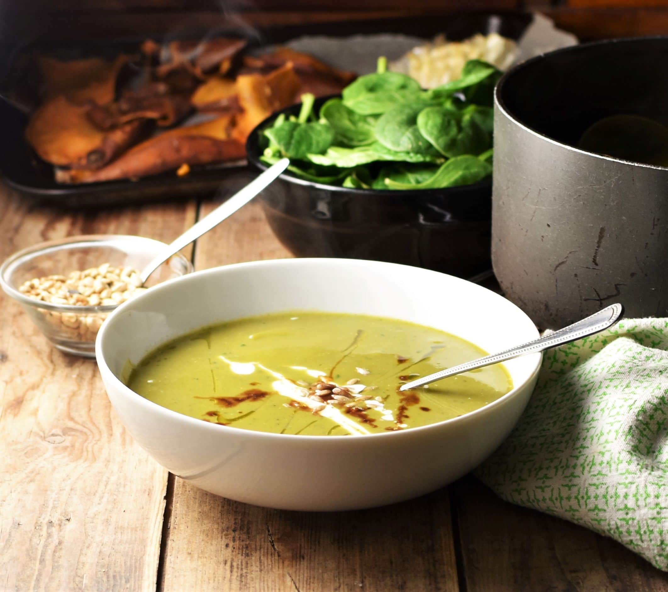 Side view of green soup in white bowl with spoon, with spinach, seeds in small dish, black pot and green cloth in background.