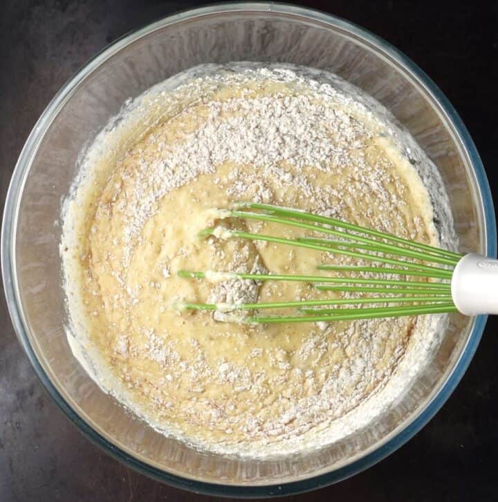 Adding flour to banana pancake batter in glass bowl with whisk.