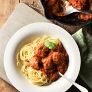 Meatballs in tomato sauce over spaghetti in bowl with fork and meatballs in sauce in pan in top right corner.