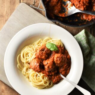 Meatballs in tomato sauce over spaghetti in bowl with fork and meatballs in sauce in pan in top right corner.