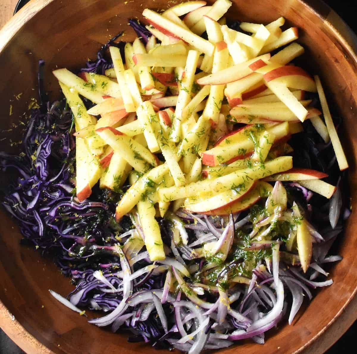 Top down view of apple matchsticks, shredded red cabbage, red onion and dressing in wooden bowl.