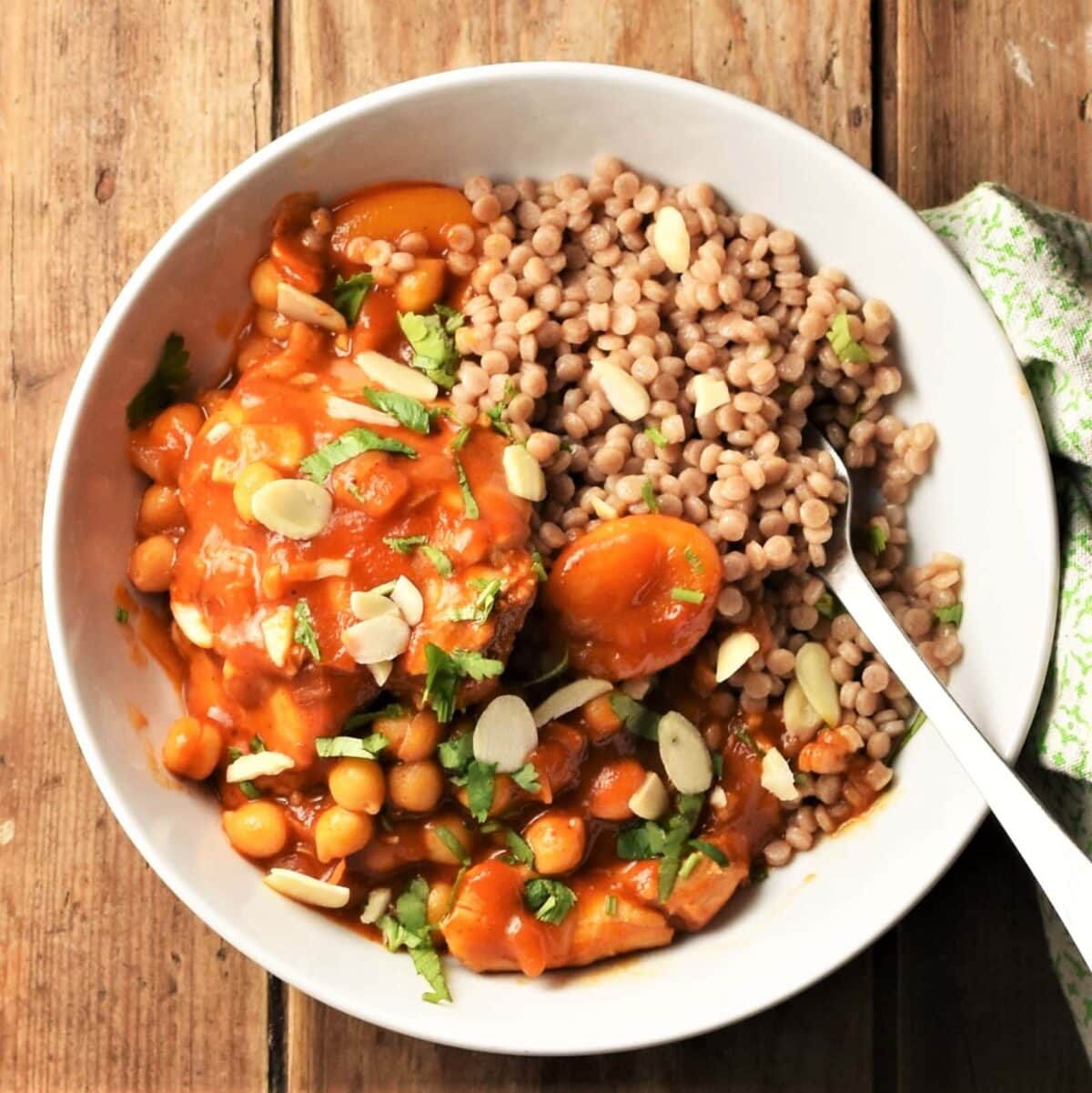 Chicken apricot in tomato sauce with giant couscous in white bowl with spoon.
