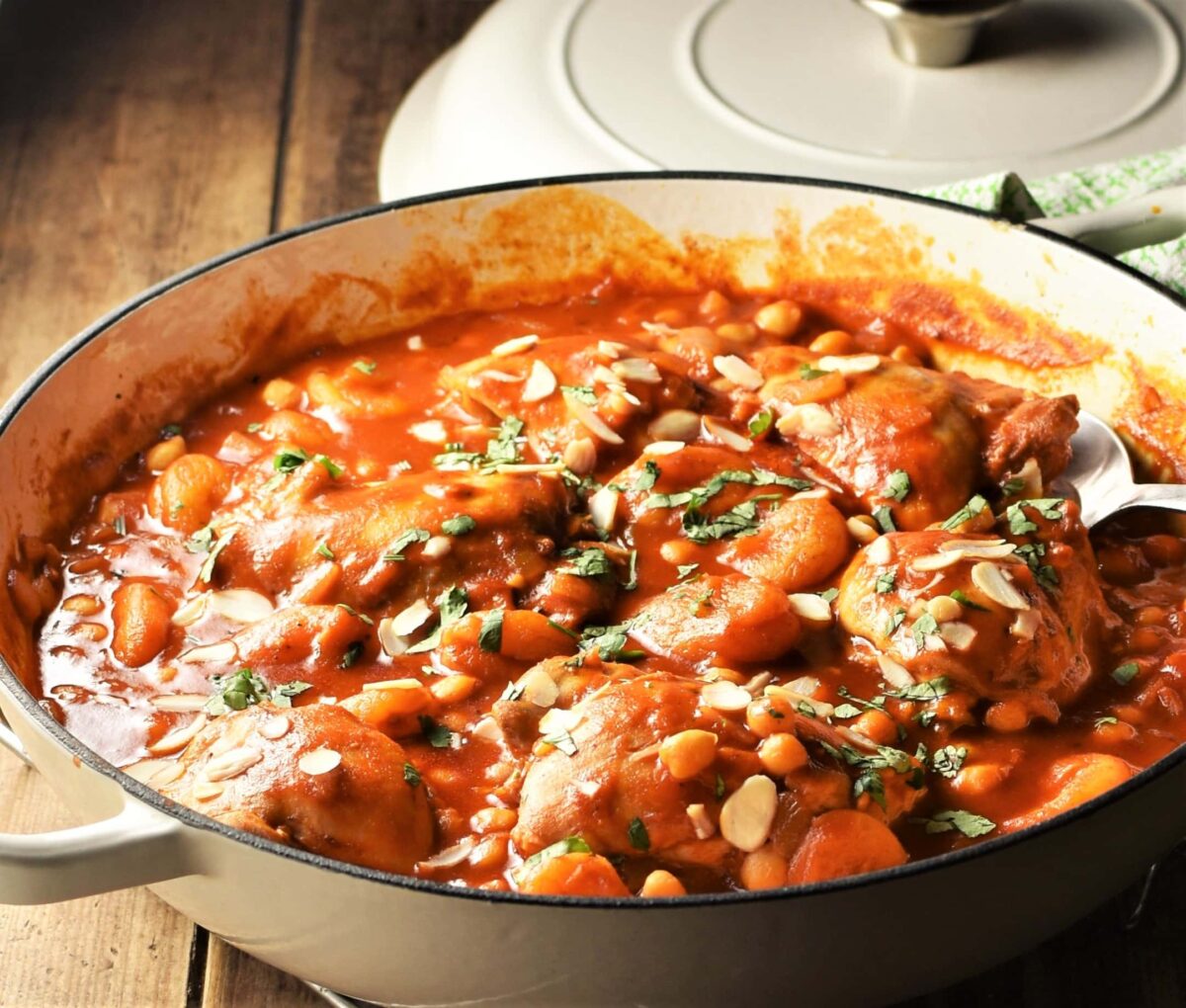 Side view of chicken thighs in tomato sauce with chickpeas in large shallow dish with lid in background.