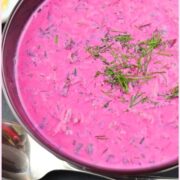 Top down view of cold beet soup with dill, spoon in front and white cloth at top.