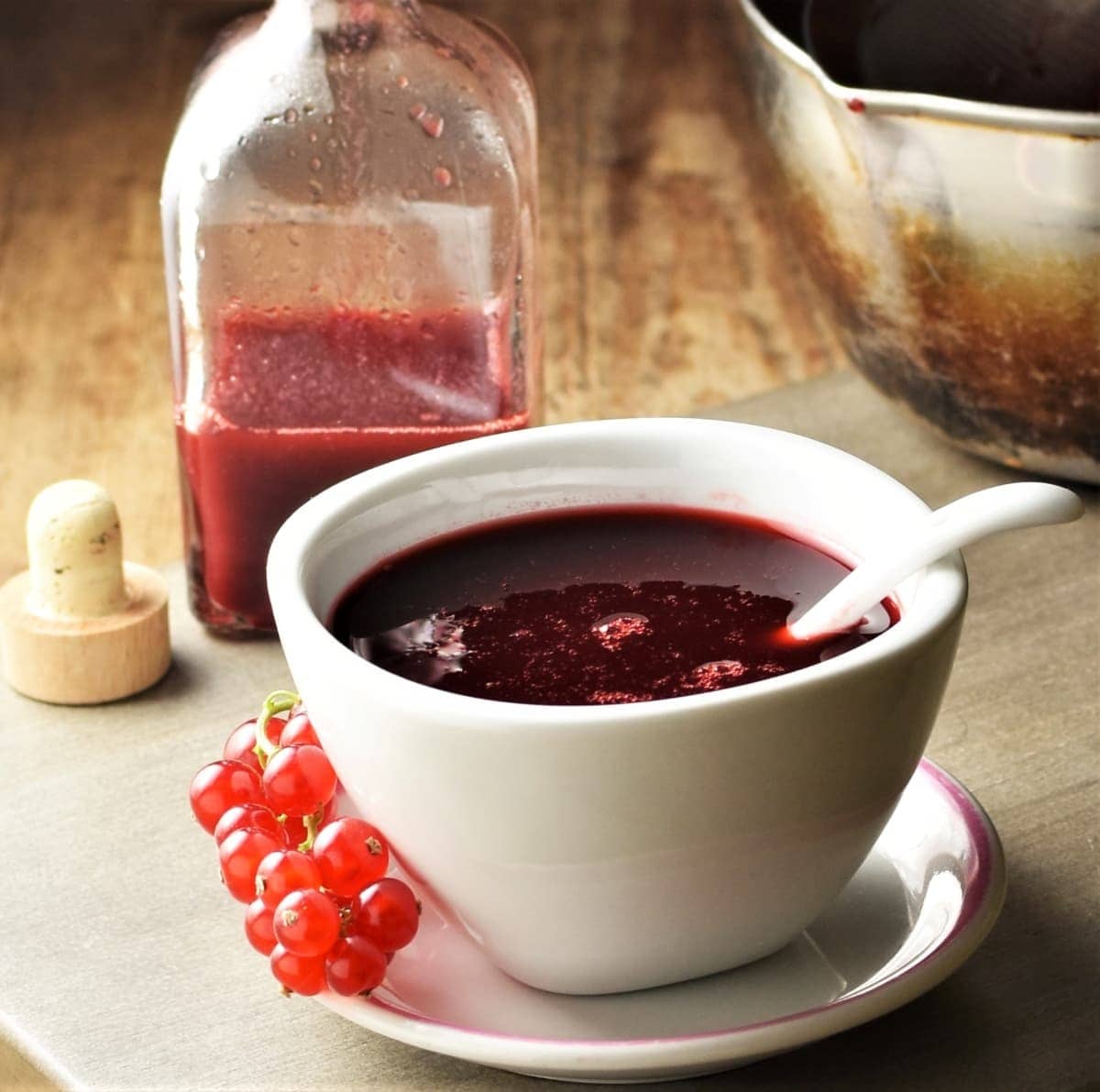 Redcurrant sauce in white bowl with spoon on top of saucer with redcurrants and sauce in bottle in background.