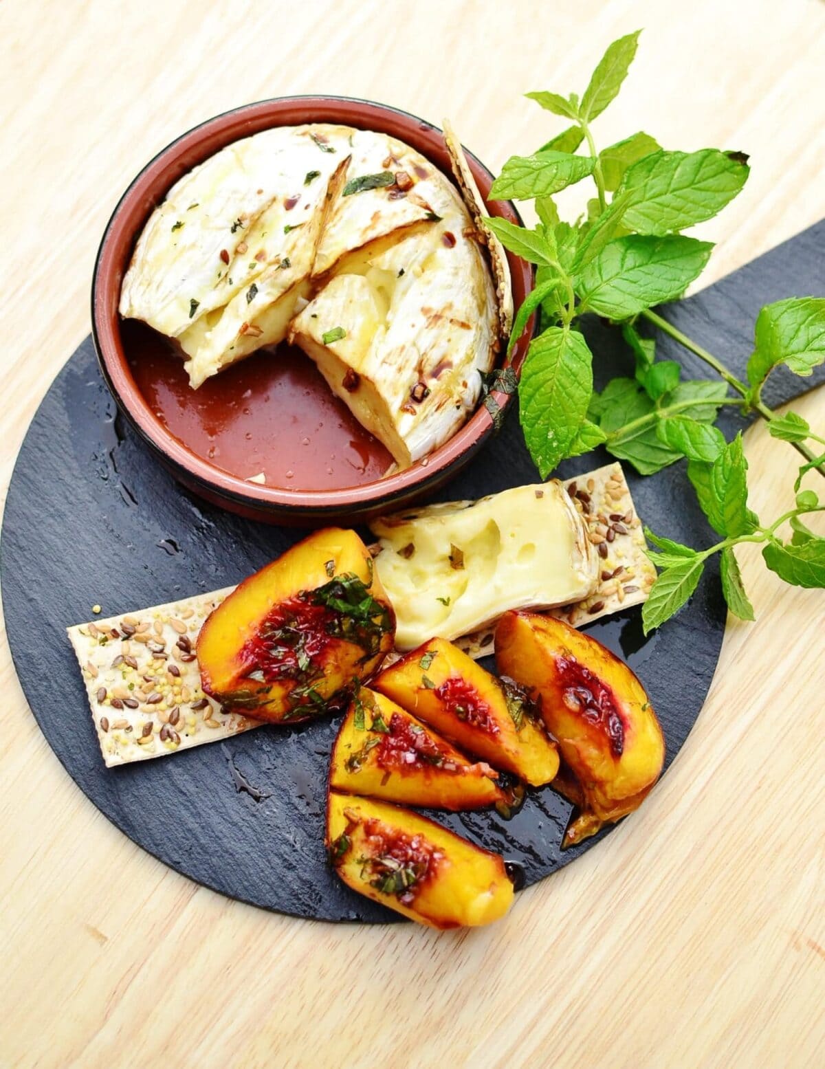 Top down view of camembert cheese inside round brown dish, with mint leaves, crackers and baked peaches pieces on top of small slate plate.